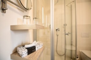 Une salle de bain avec une douche à parois de verre, une étagère en bois sur laquelle repose une boîte de mouchoirs, et un miroir au-dessus de l’étagère.