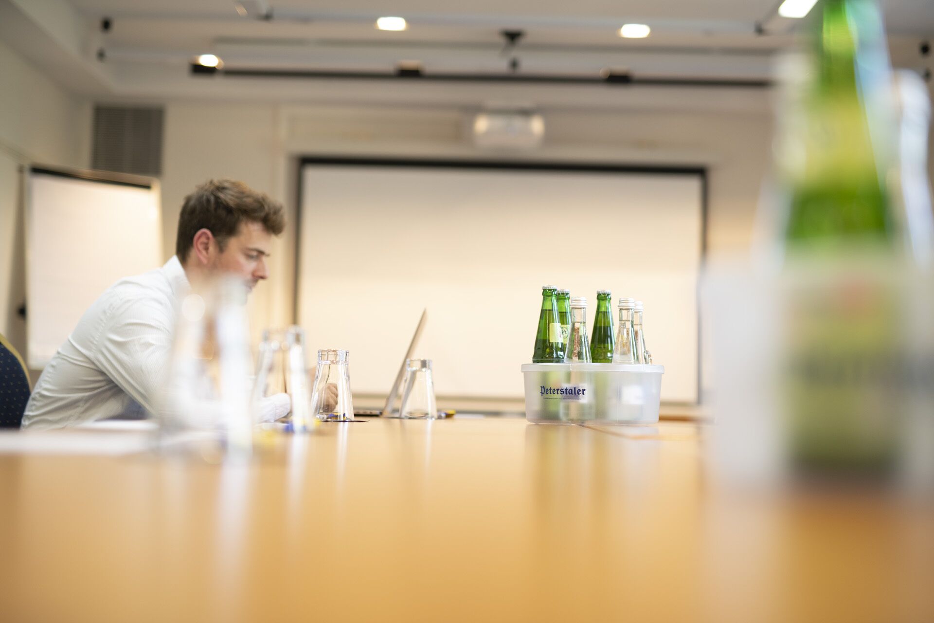 A conference room with a table holding several bottles and glasses, a projector screen, and a person seated at the table.