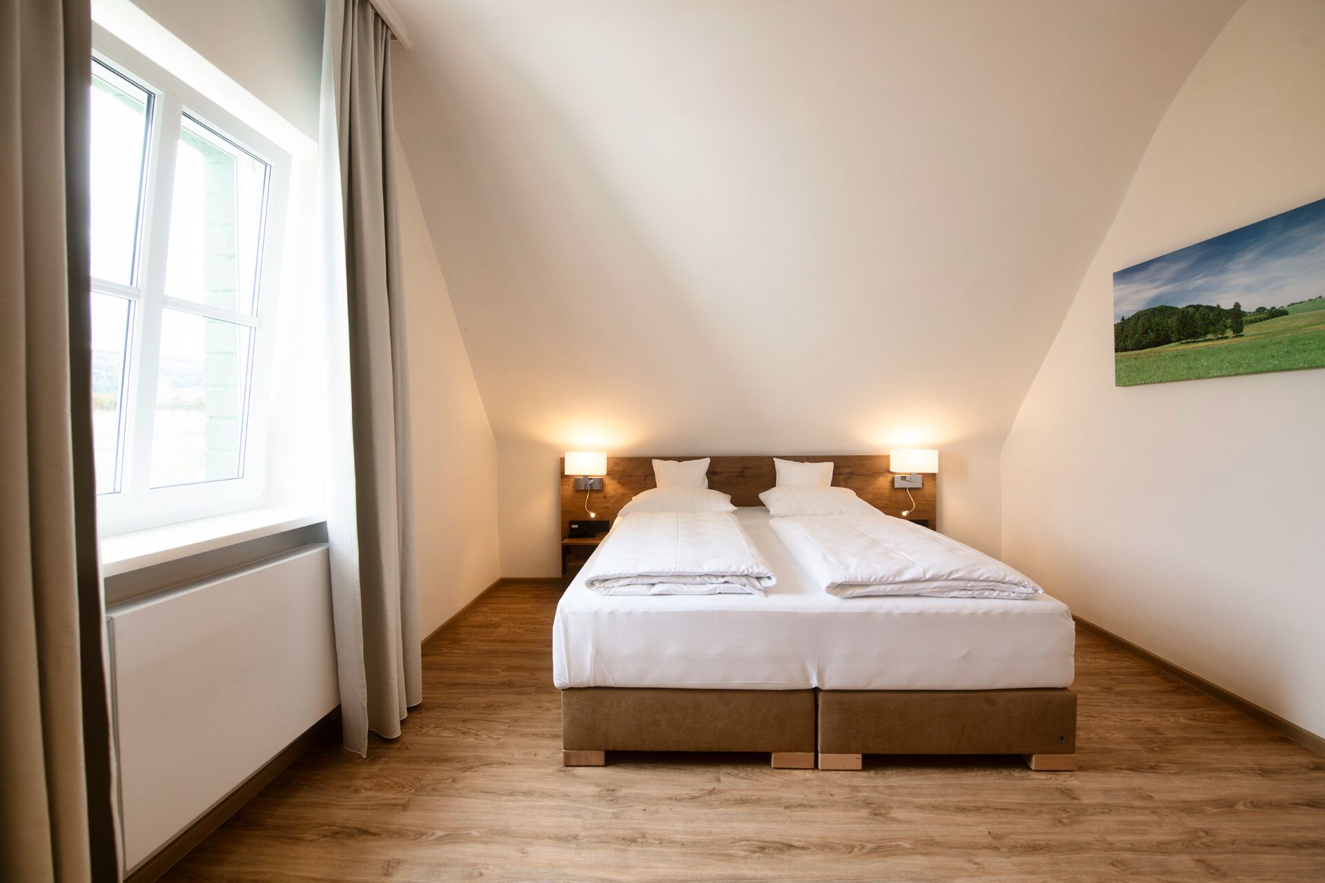 Un dormitorio con cama, piso de madera, dos lámparas de noche, una ventana con cortinas y un cuadro en la pared.