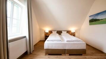 Un dormitorio con cama, piso de madera, dos lámparas de noche, una ventana con cortinas y un cuadro en la pared.