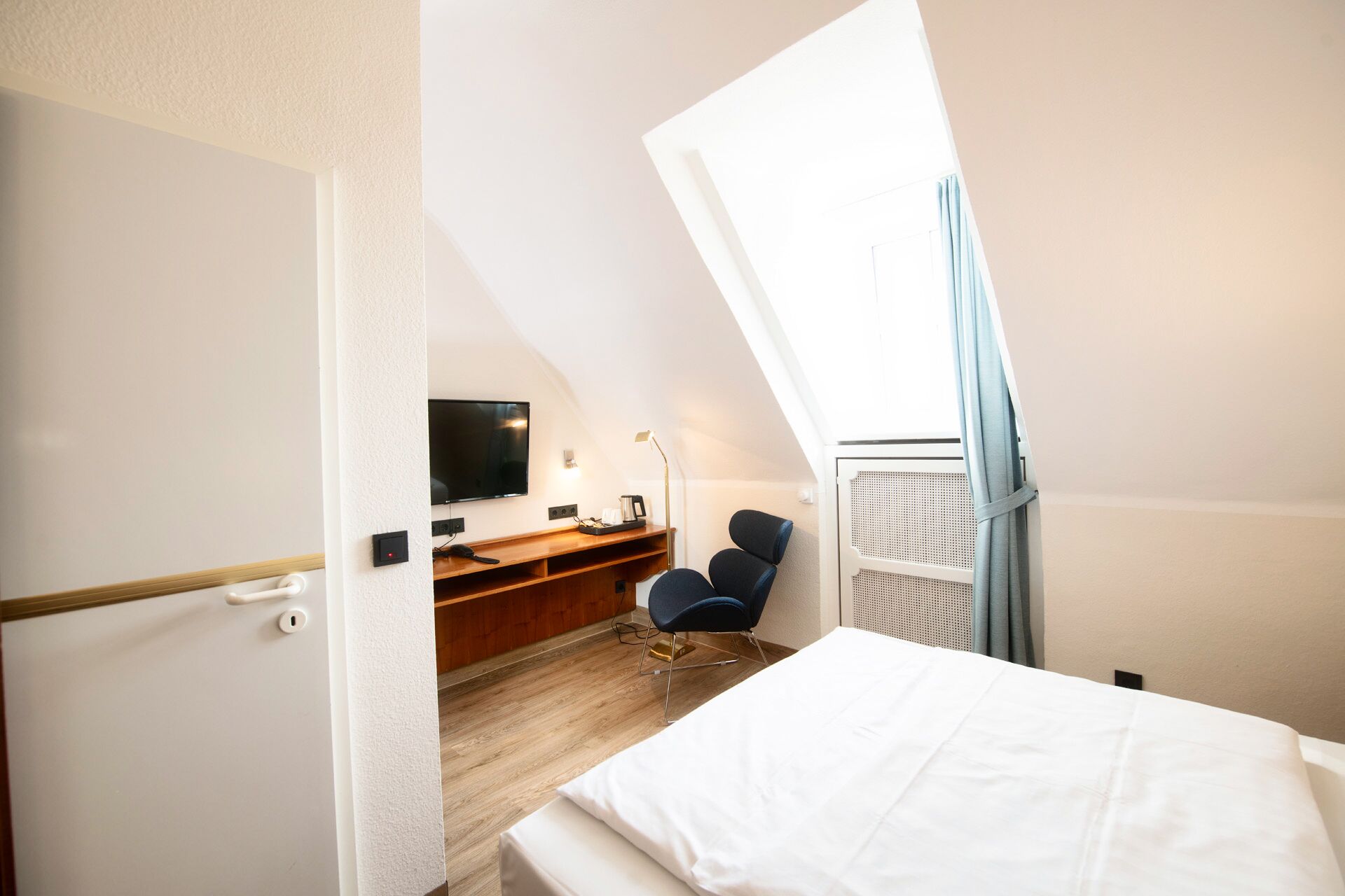A small, well-lit room with a bed, a desk, a chair, and a TV mounted on the wall.