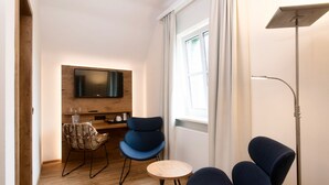 Ein modernes Wohnzimmer mit einem an einer Holzwand montierten Flachbildfernseher, zwei blauen Sesseln, einem kleinen runden Holztisch, einem Korbstuhl und einer Stehlampe.