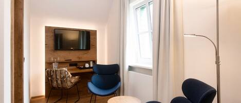 Una sala de estar moderna con un televisor de pantalla plana montado en una pared de madera, dos sillones azules, una mesita redonda de madera, una silla de mimbre y una lámpara de pie.