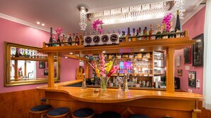 Zona de bar con mostrador de madera, varios taburetes, un espejo y una variedad de botellas y vasos en estantes.