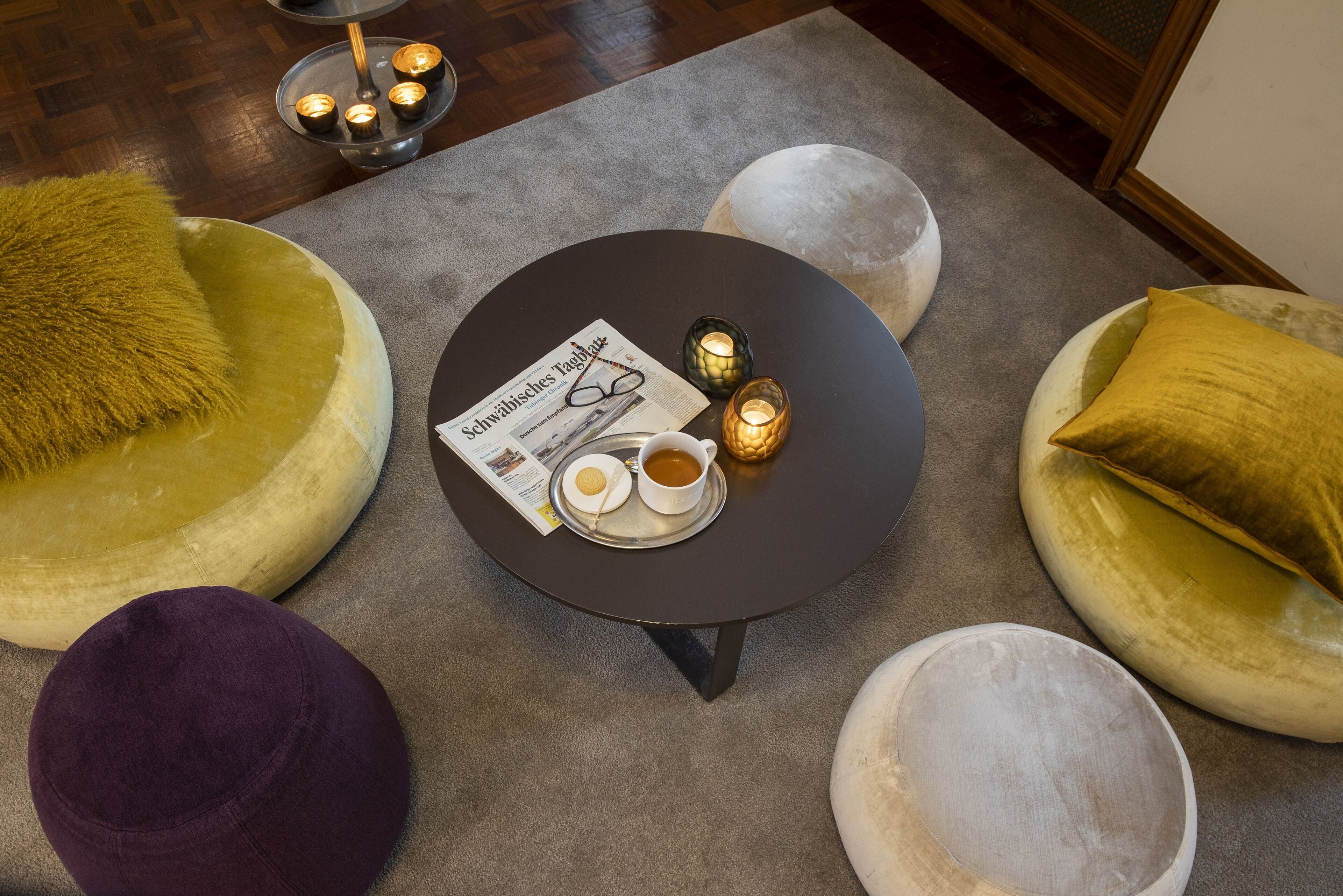 A round table with a newspaper, a cup of coffee, and a plate with a cookie. There are candles on the table and a fluffy pillow on the floor.
