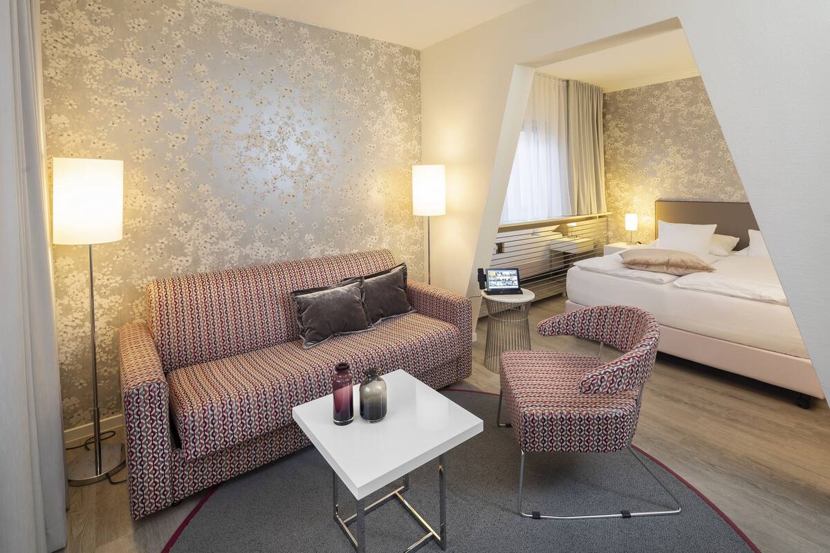 A hotel room with a patterned sofa, a small white table, a patterned armchair, a bed, and a bedside table with a lamp.