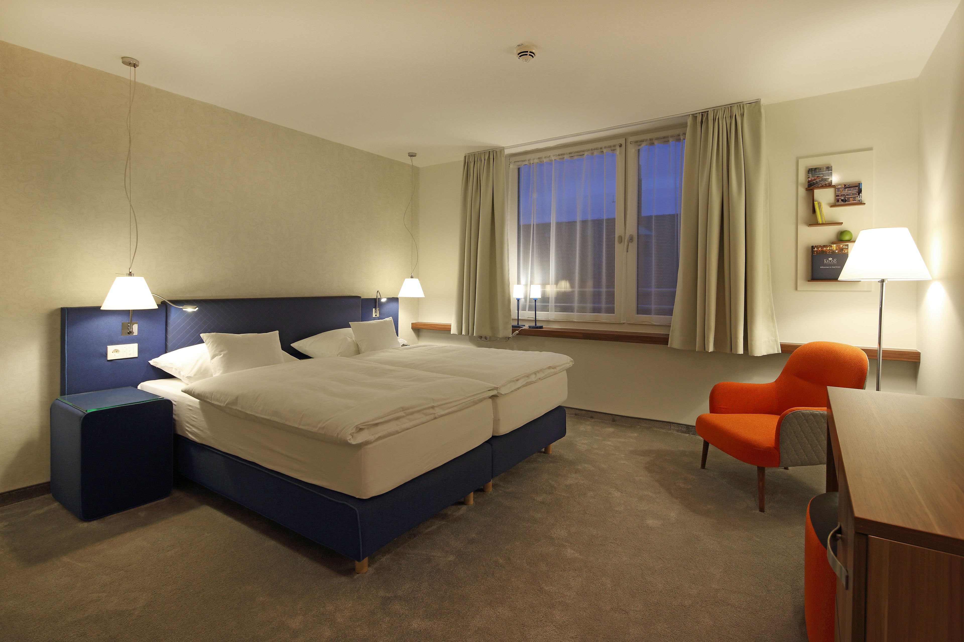 Une chambre d’hôtel avec un lit, deux tables de chevet, un bureau, une chaise orange et une fenêtre avec des rideaux.
