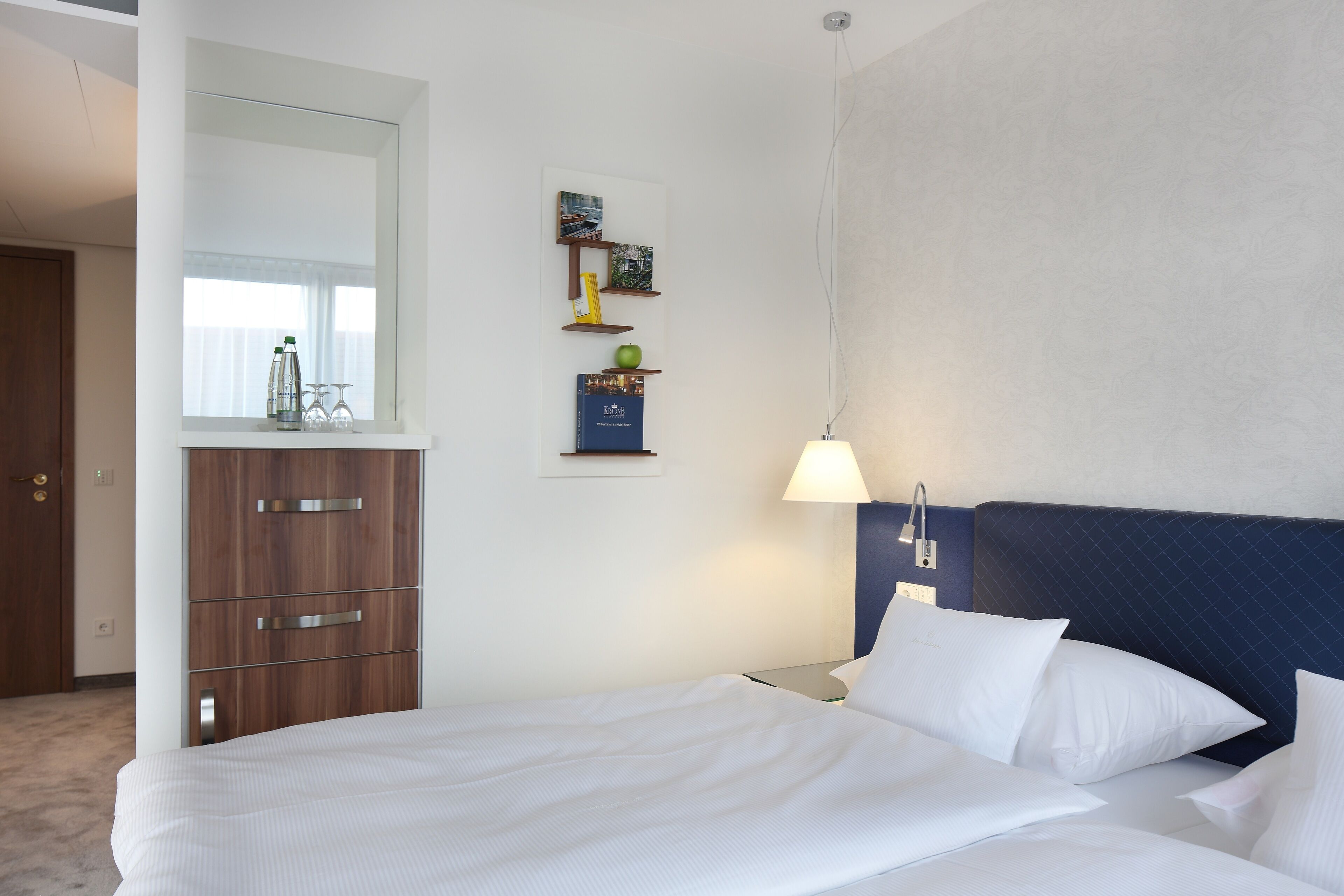 Ein Hotelzimmer mit einem Bett, einem Nachttisch, einer Lampe, einem Schrank und einem an der Wand befestigten Regal mit Dekoration.