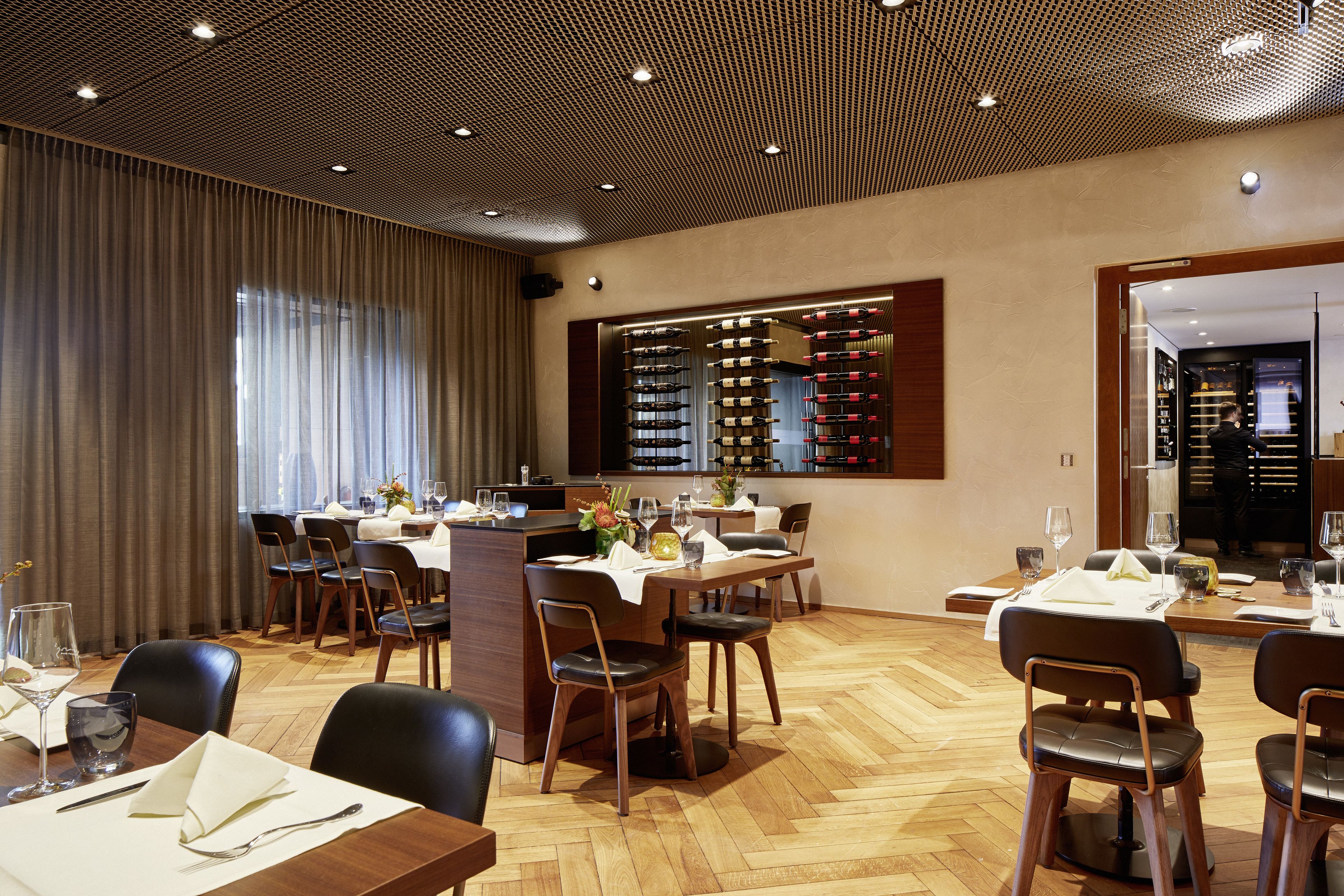 Un restaurante con suelo de madera, mesas dispuestas con manteles blancos y una estantería con botellas de vino al fondo.