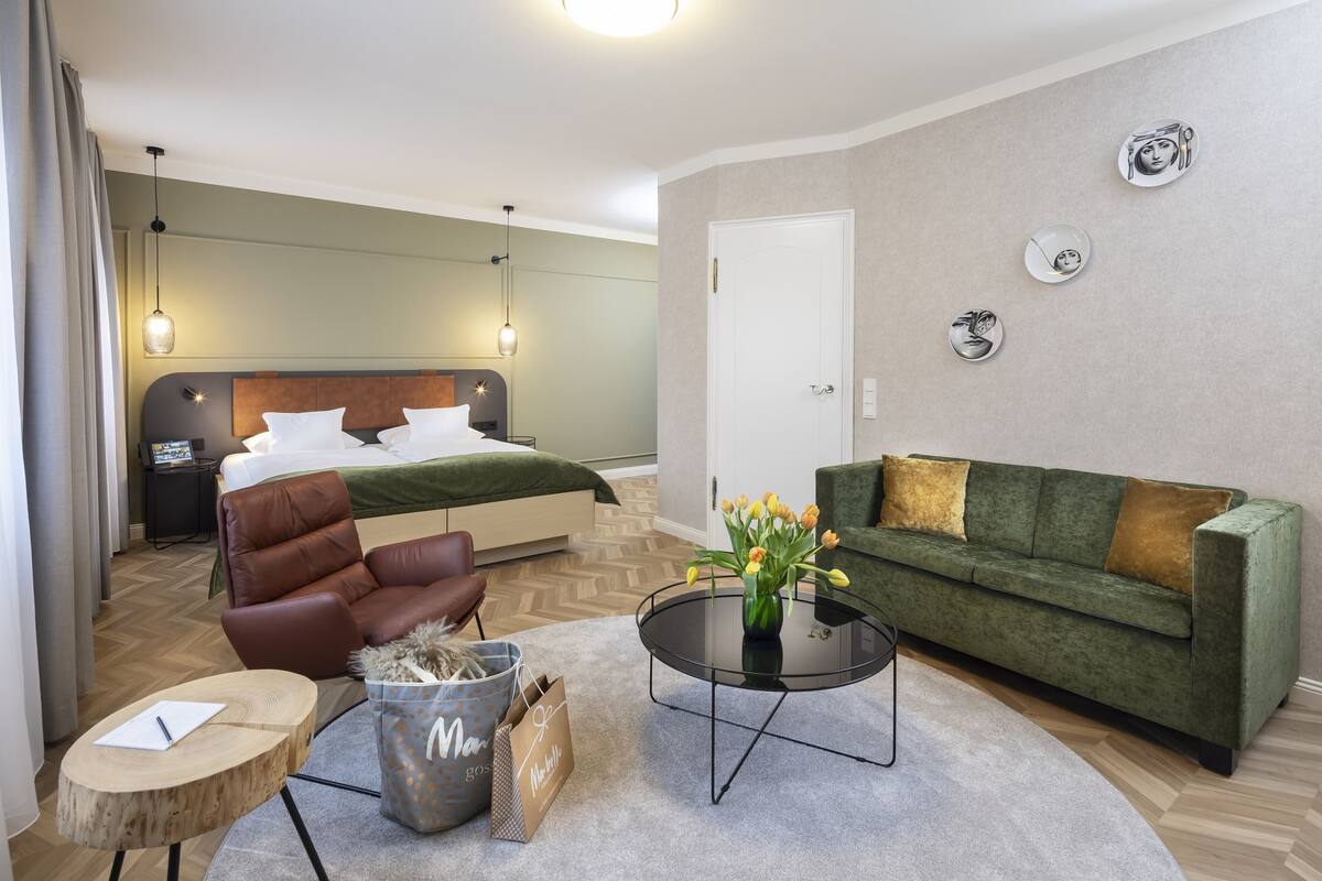 A modern hotel room with a bed, a green sofa, a coffee table, a brown leather chair, and a small side table.