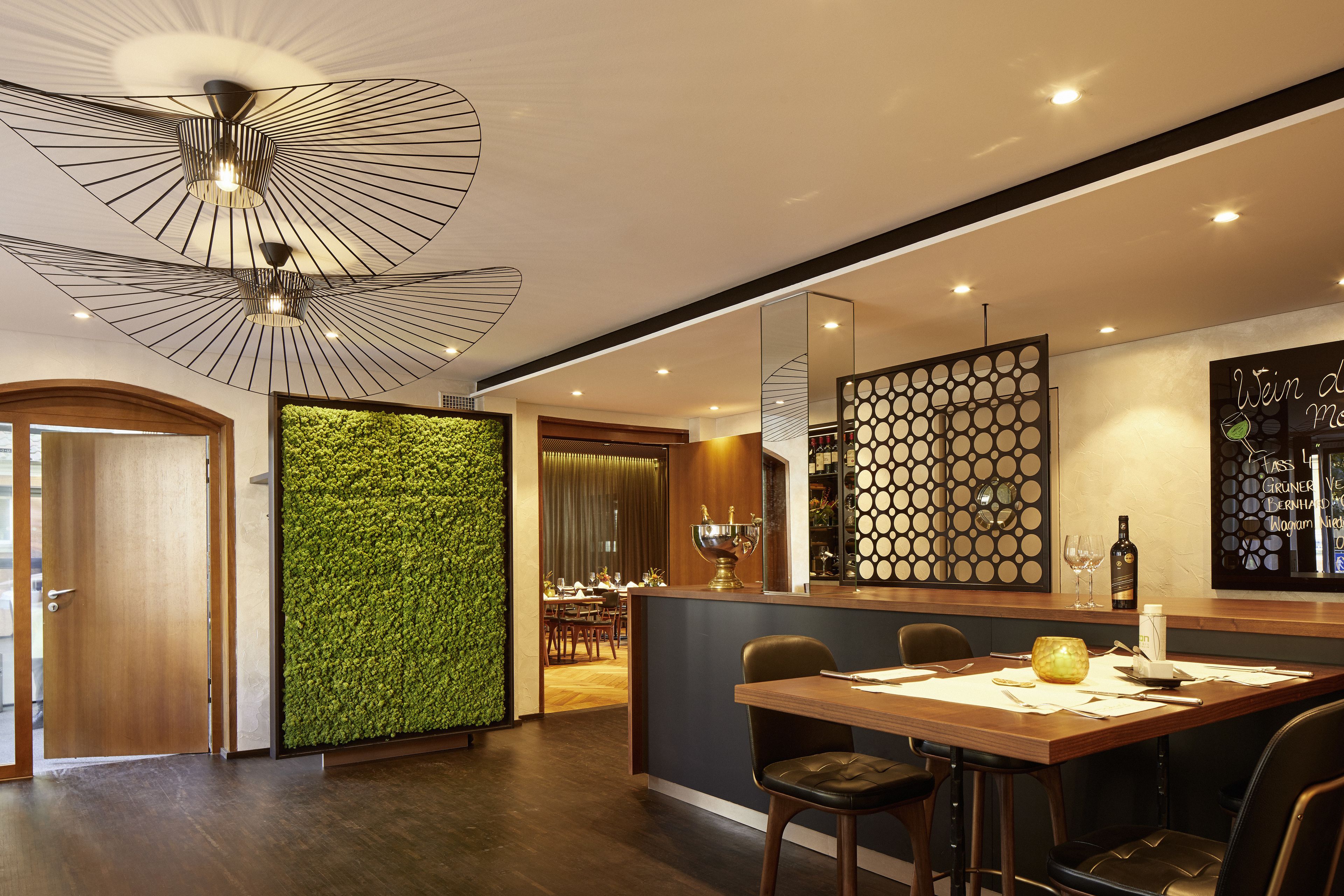 A modern dining area with a wooden table, chairs, a green vertical garden wall, and a ceiling with a geometric light fixture.
