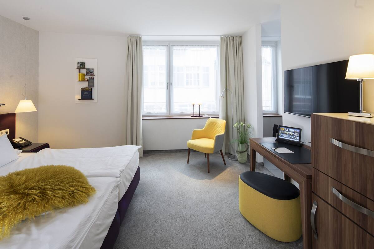 A hotel room with a bed, a desk with a TV, a chair, and a small table with a plant.