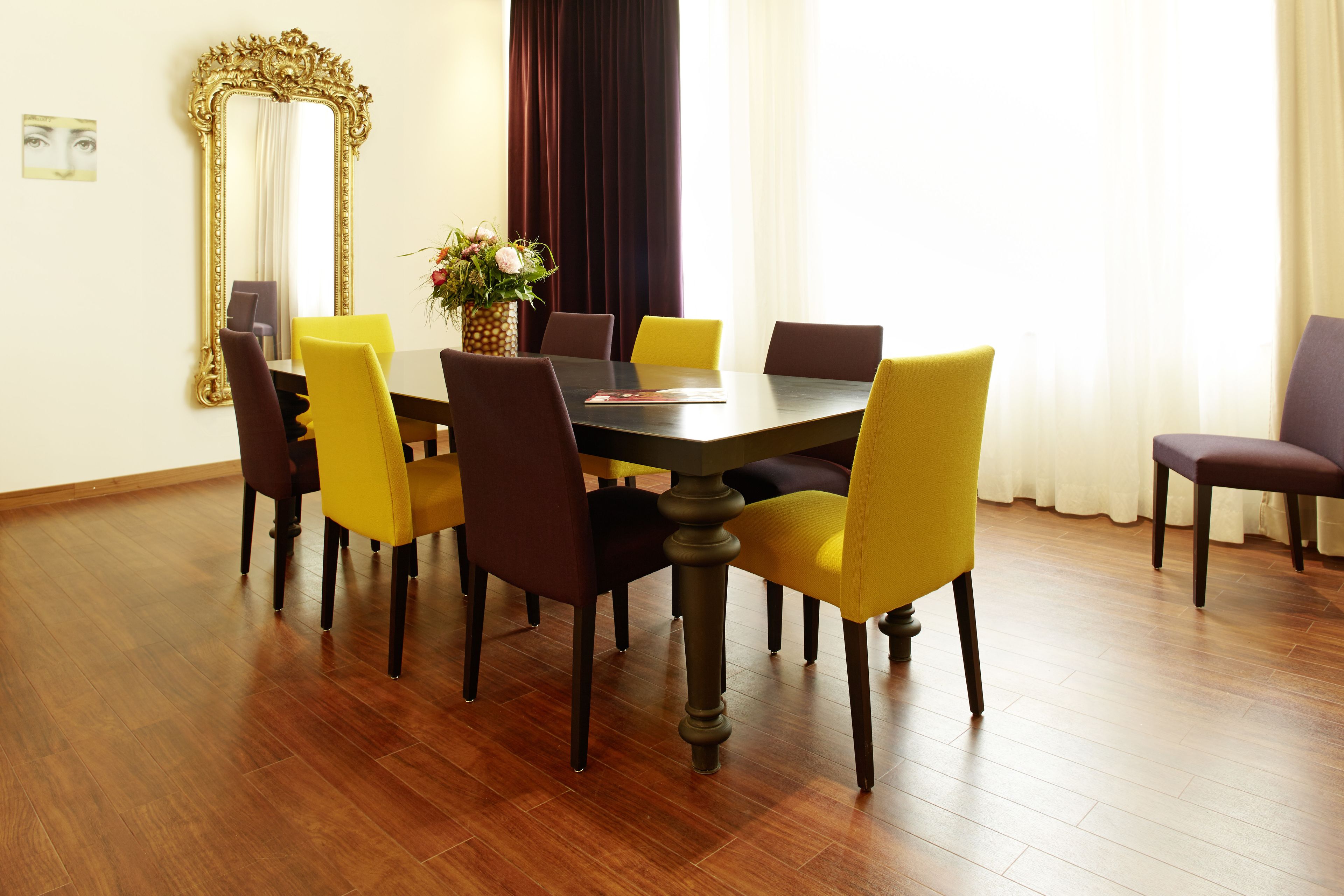 A dining area with a wooden table and chairs, a large mirror, and a vase with flowers.