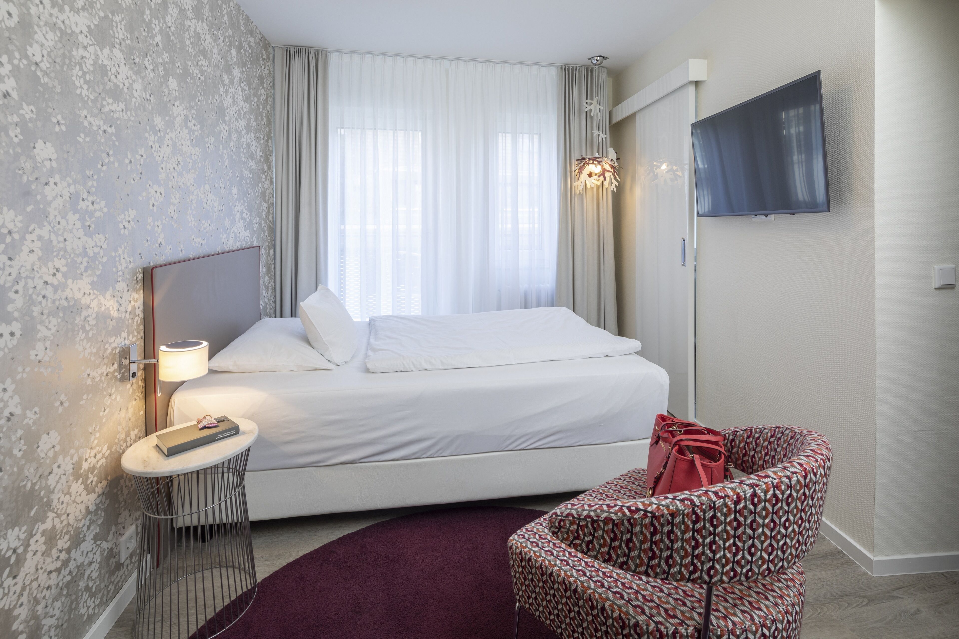 A hotel room with a bed, a round side table with a lamp, a TV mounted on the wall, and a patterned armchair.