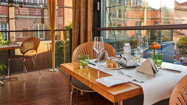 Un área de comedor con una mesa de madera dispuesta para dos personas, que incluye un vaso de agua, una copa de vino, un plato con un croissant y un florero con una flor.