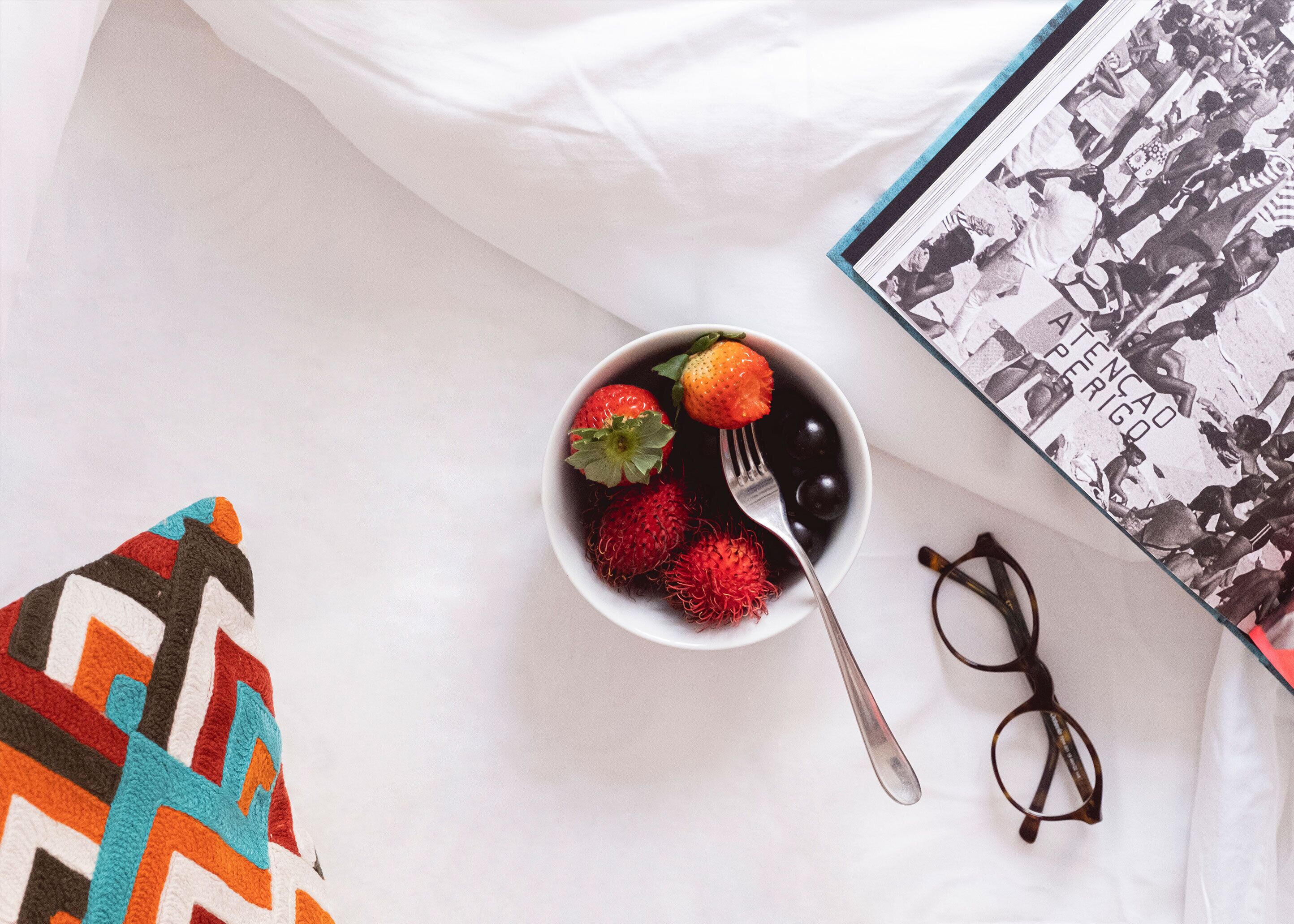 A bowl of fruit with strawberries and rambutan on a white surface, a pair of glasses, and a book with a black and white cover.