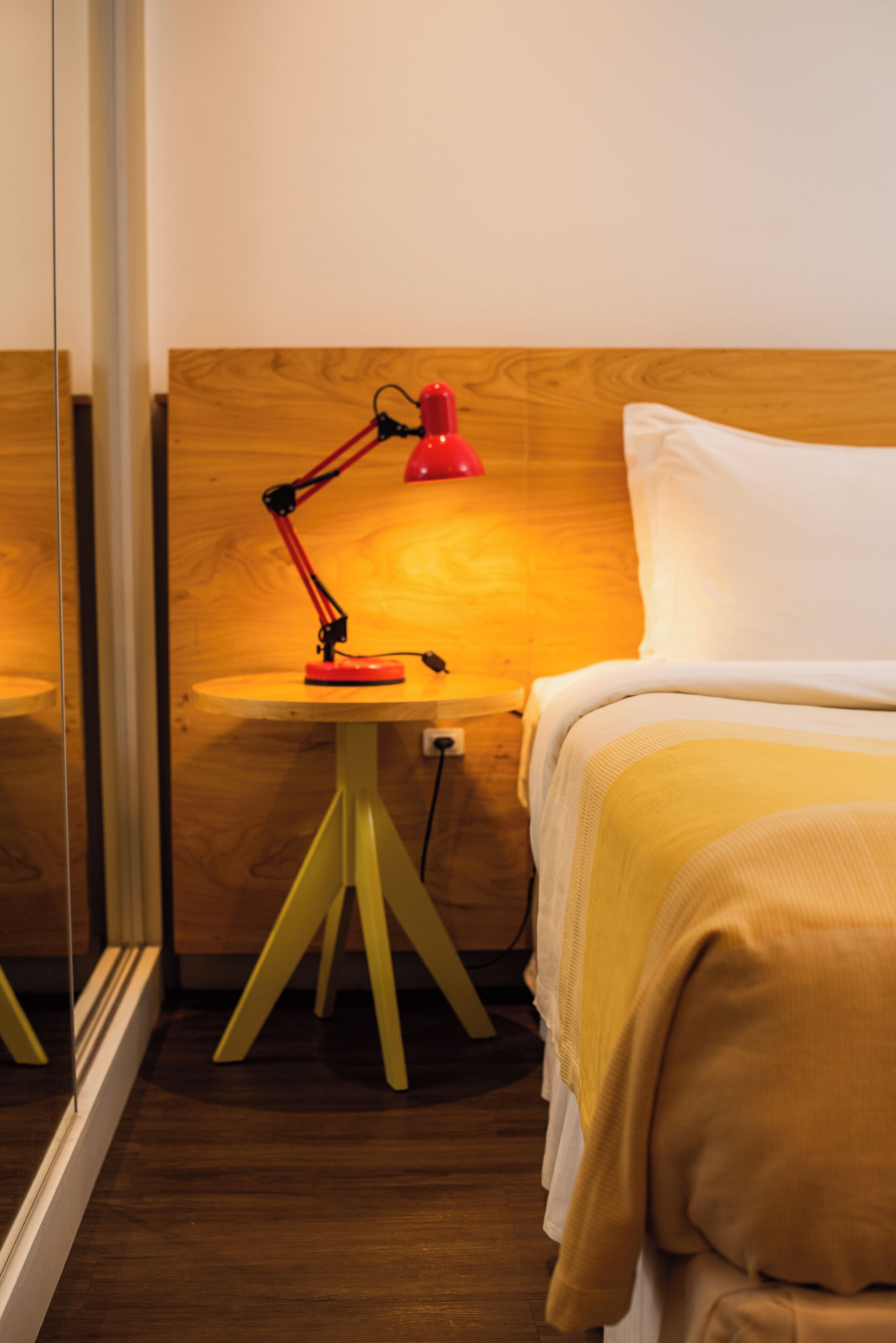 A neatly made bed with a white pillow and a yellow blanket, a red desk lamp on a small wooden table, and a wooden headboard.