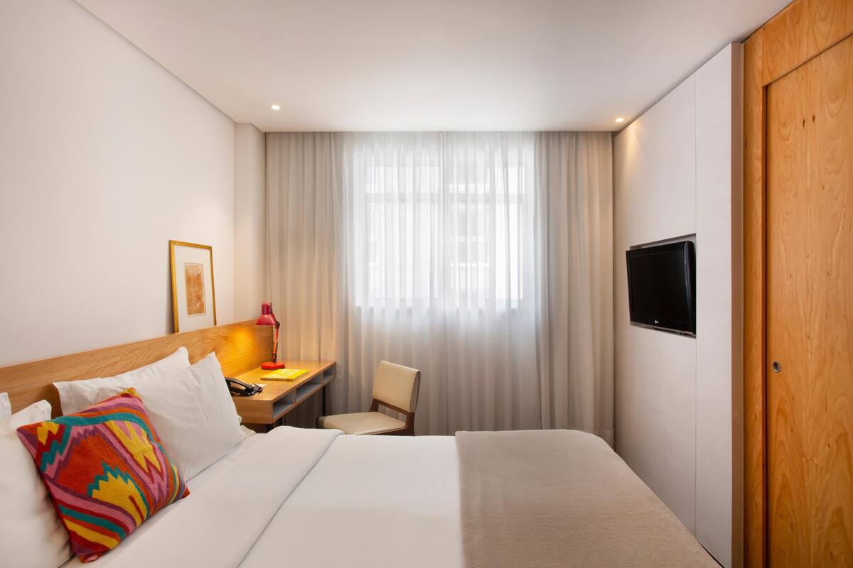 Habitación de hotel con cama, escritorio, silla, televisor y ventana con cortinas.