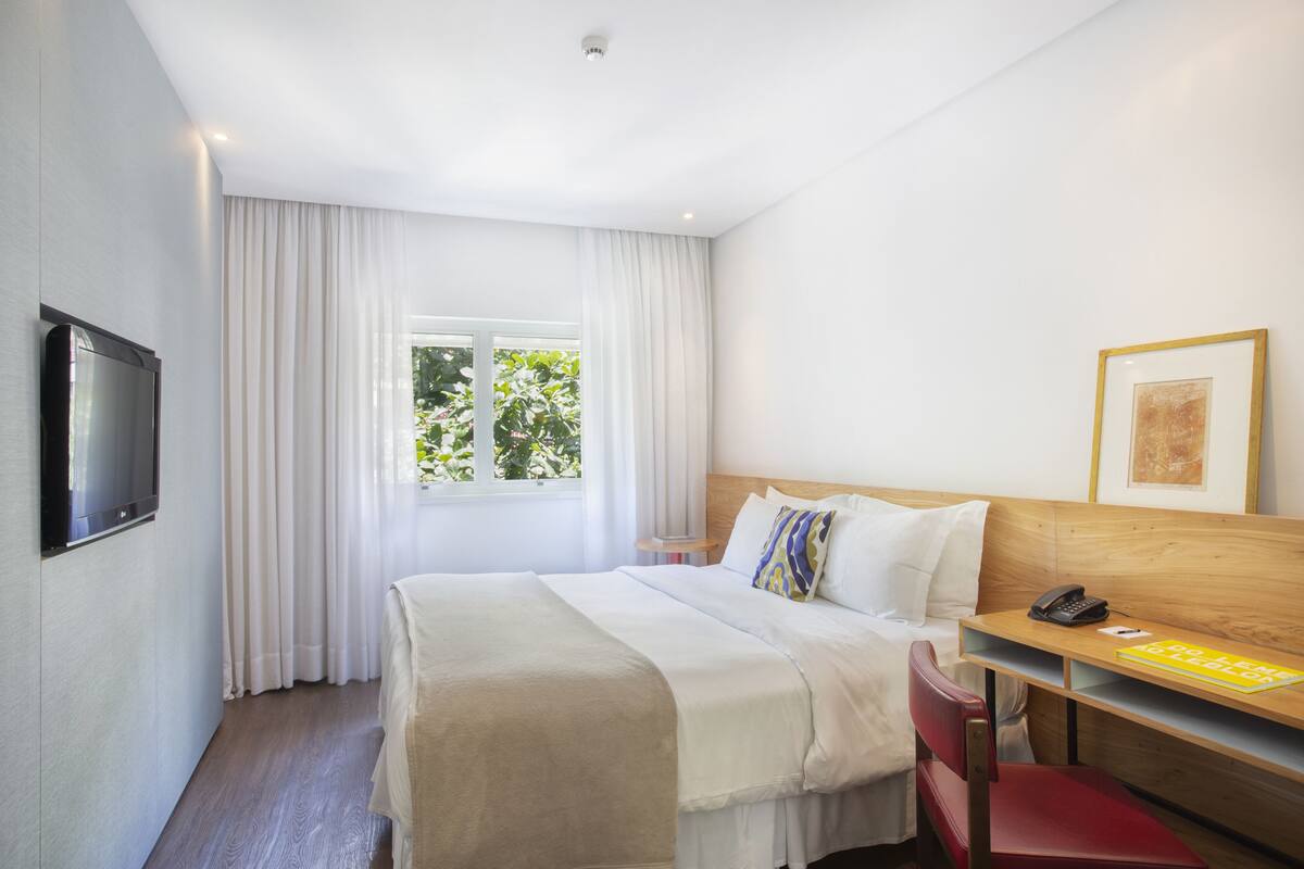 Una habitación de hotel con cama, escritorio con teléfono, silla, televisión y ventana con cortinas.