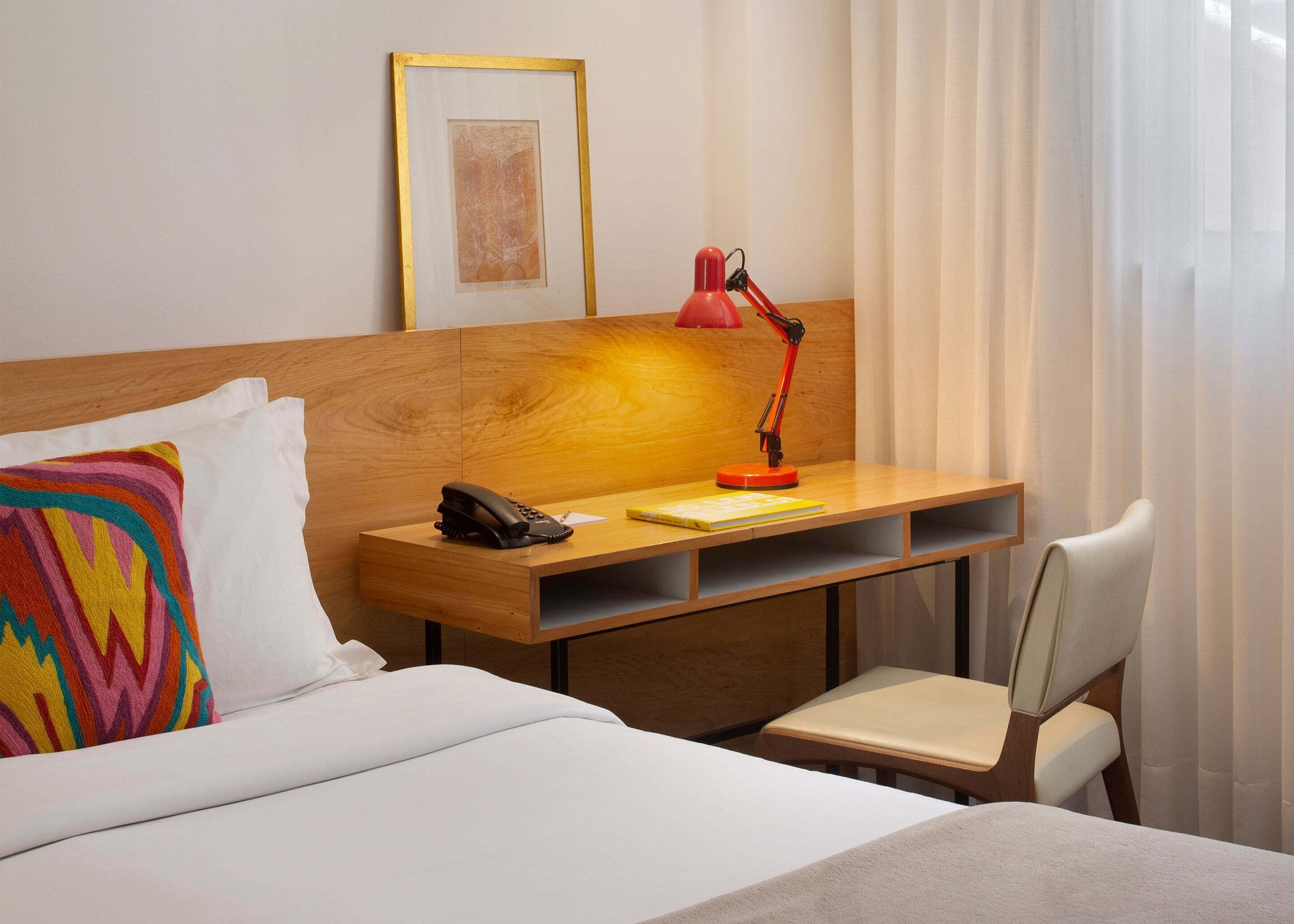 A hotel room with a bed, a desk with a telephone, a red desk lamp, a chair, and a framed picture on the wall.