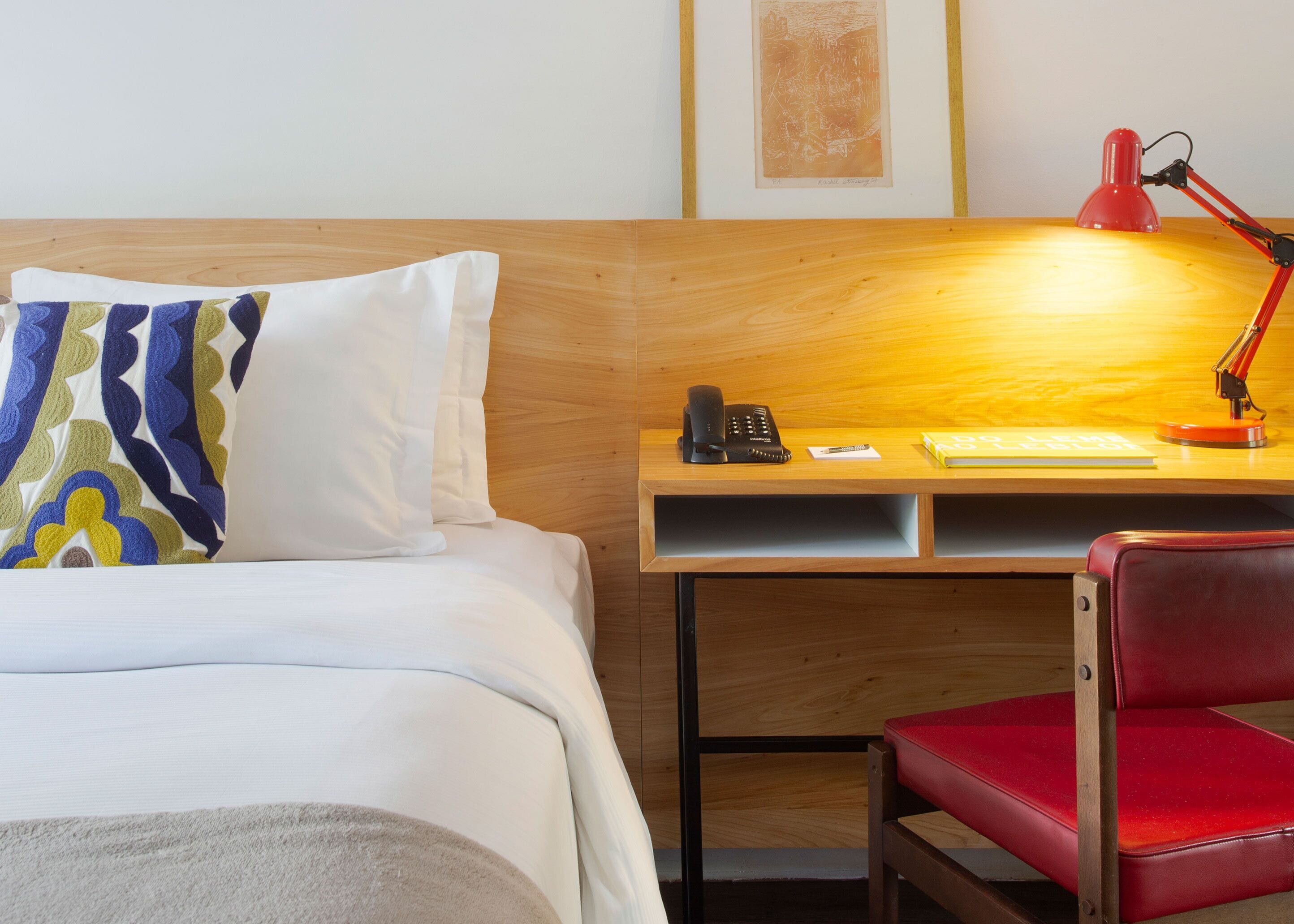 Ein Hotelzimmer mit einem Bett, einem Schreibtisch mit Telefon, einem roten Sessel und einer roten Schreibtischlampe.