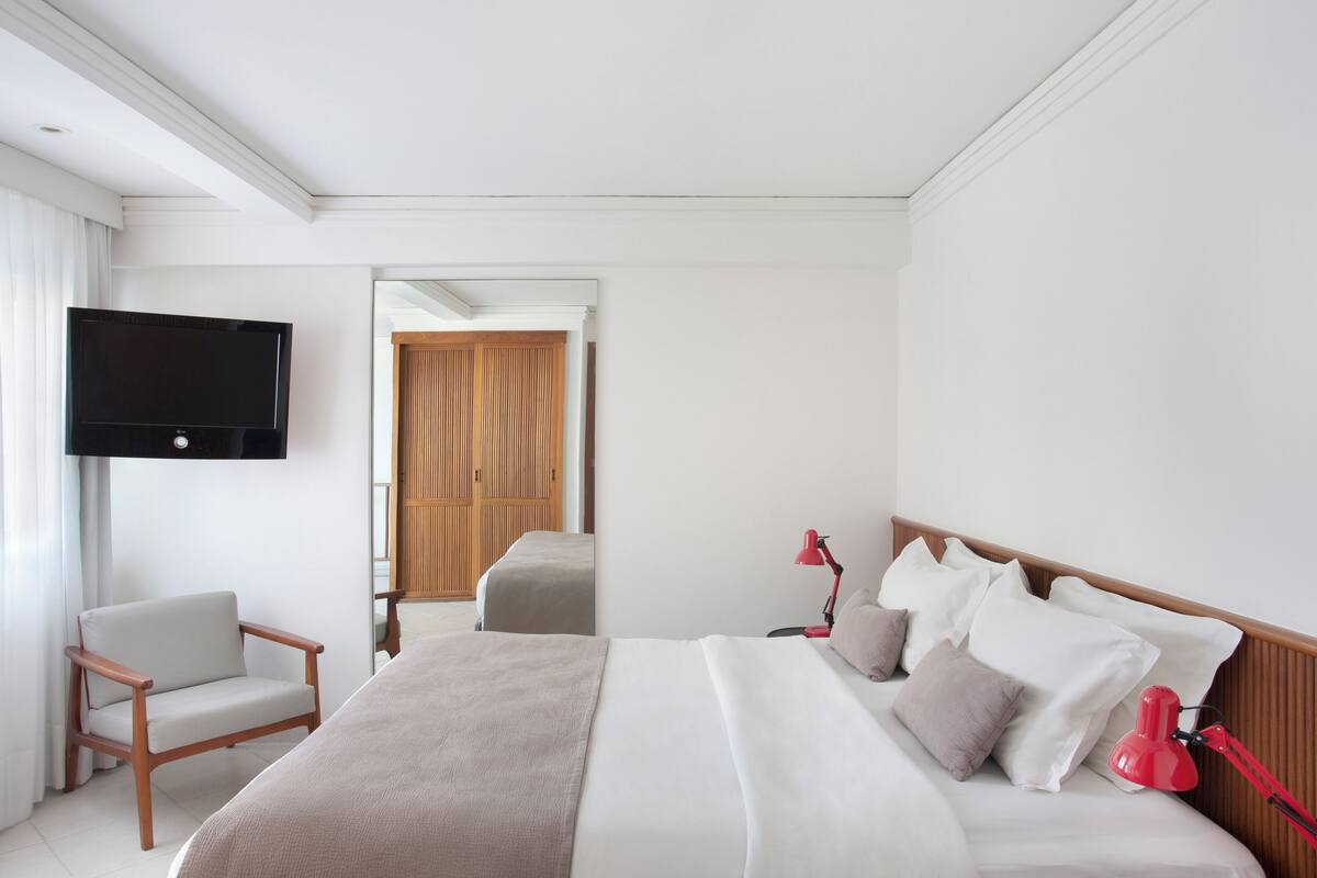 Una habitación de hotel con cama, televisión montada en la pared, una silla y una lámpara de escritorio.