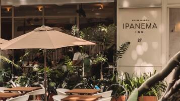 Uma área de estar externa com mesas e cadeiras, cercada por plantas em vasos. Uma placa com 'IPANEMA INN' é visível no edifício.
