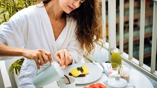 Una persona sirve café en una taza en un balcón con una mesa, fruta y vistas a edificios.