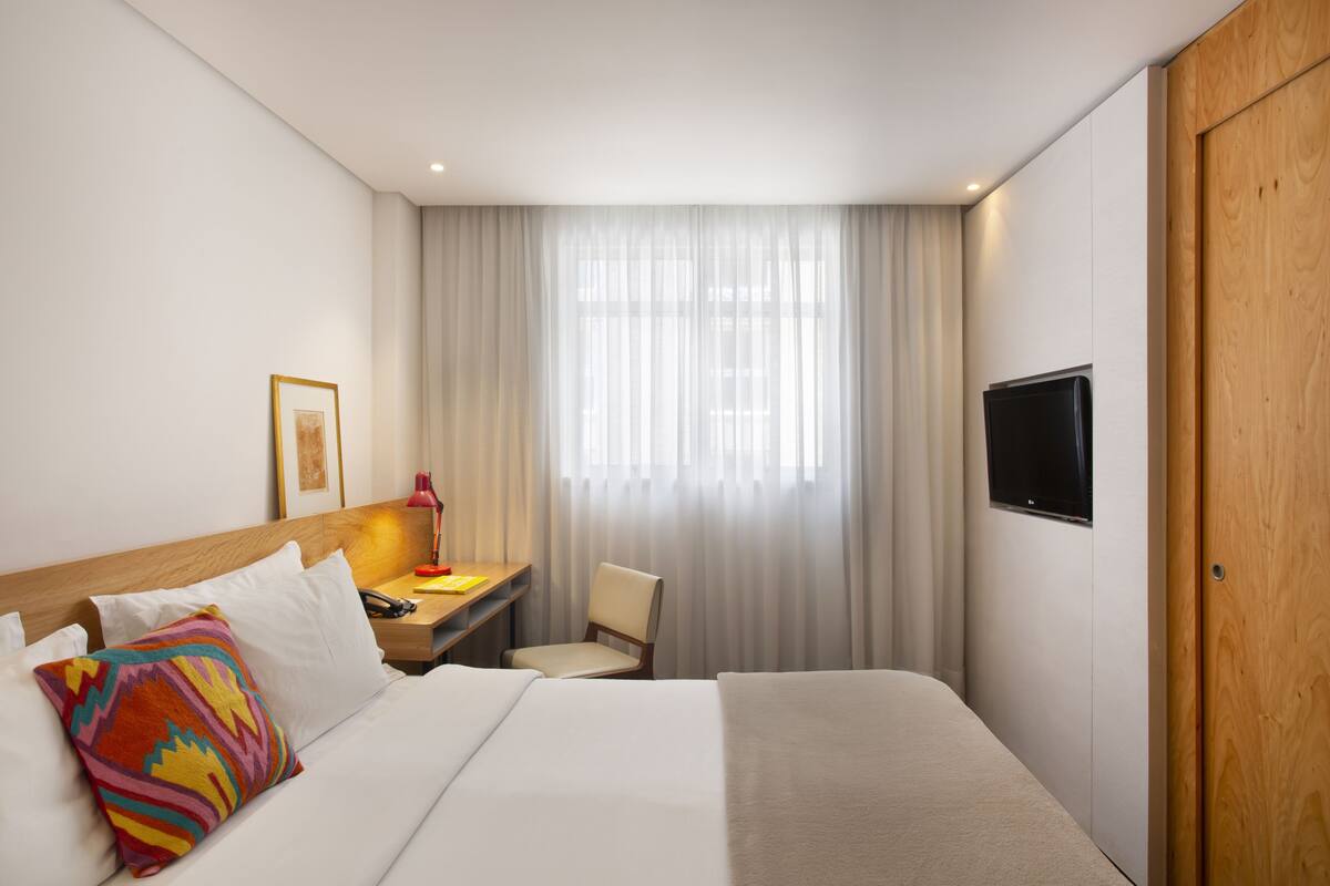 Una habitación de hotel con cama, escritorio, silla, televisión y ventana con cortinas.