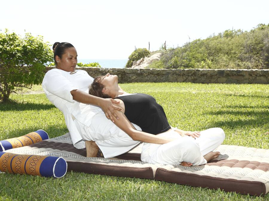 A person performing a yoga pose on a mat outdoors with grass and a stone wall in the background.