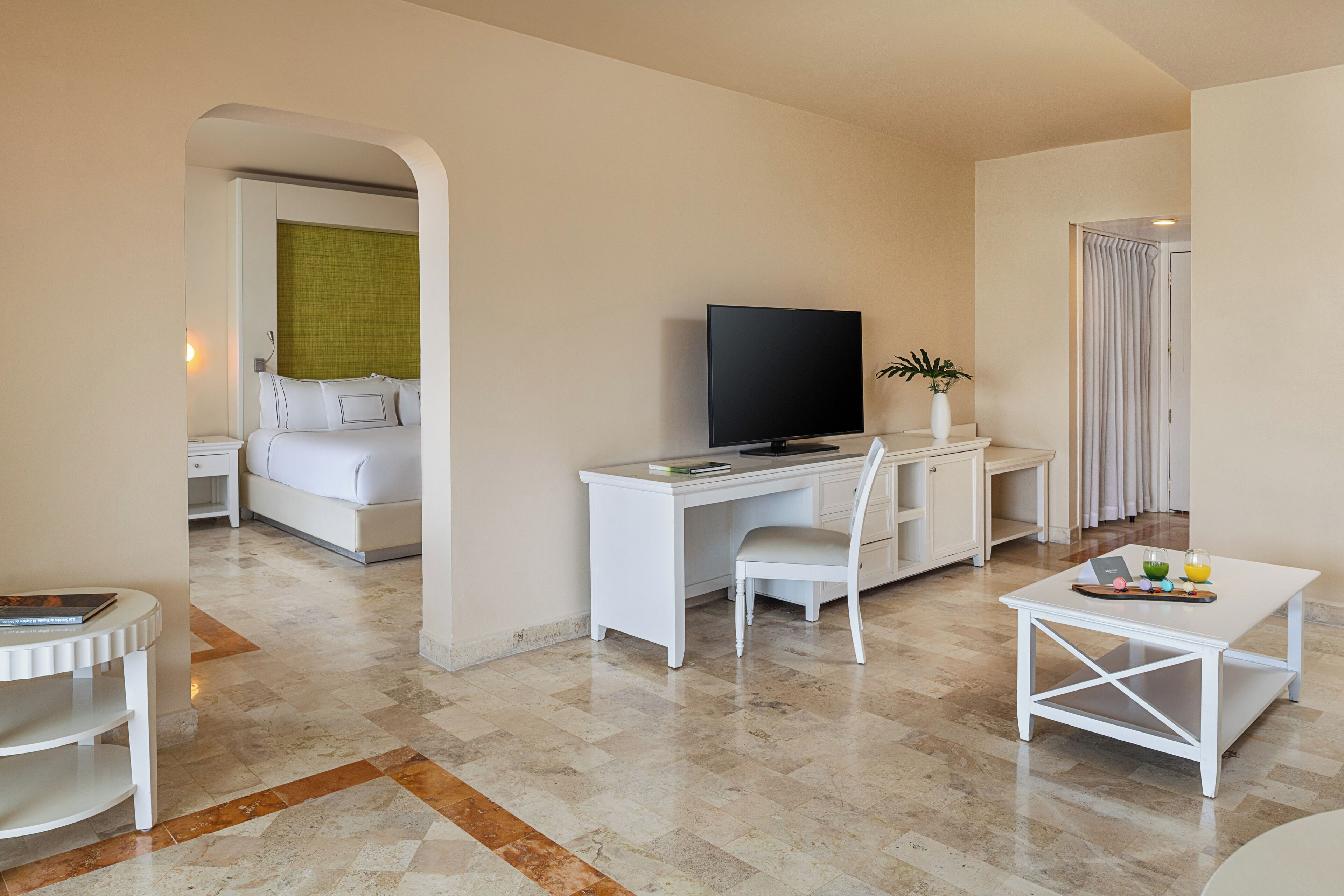 A hotel room with a bed, a desk, a television, a chair, a small table with a tray, and a plant.