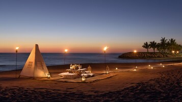Escena de playa con una carpa, mobiliario para comer y un promontorio rocoso con palmeras.