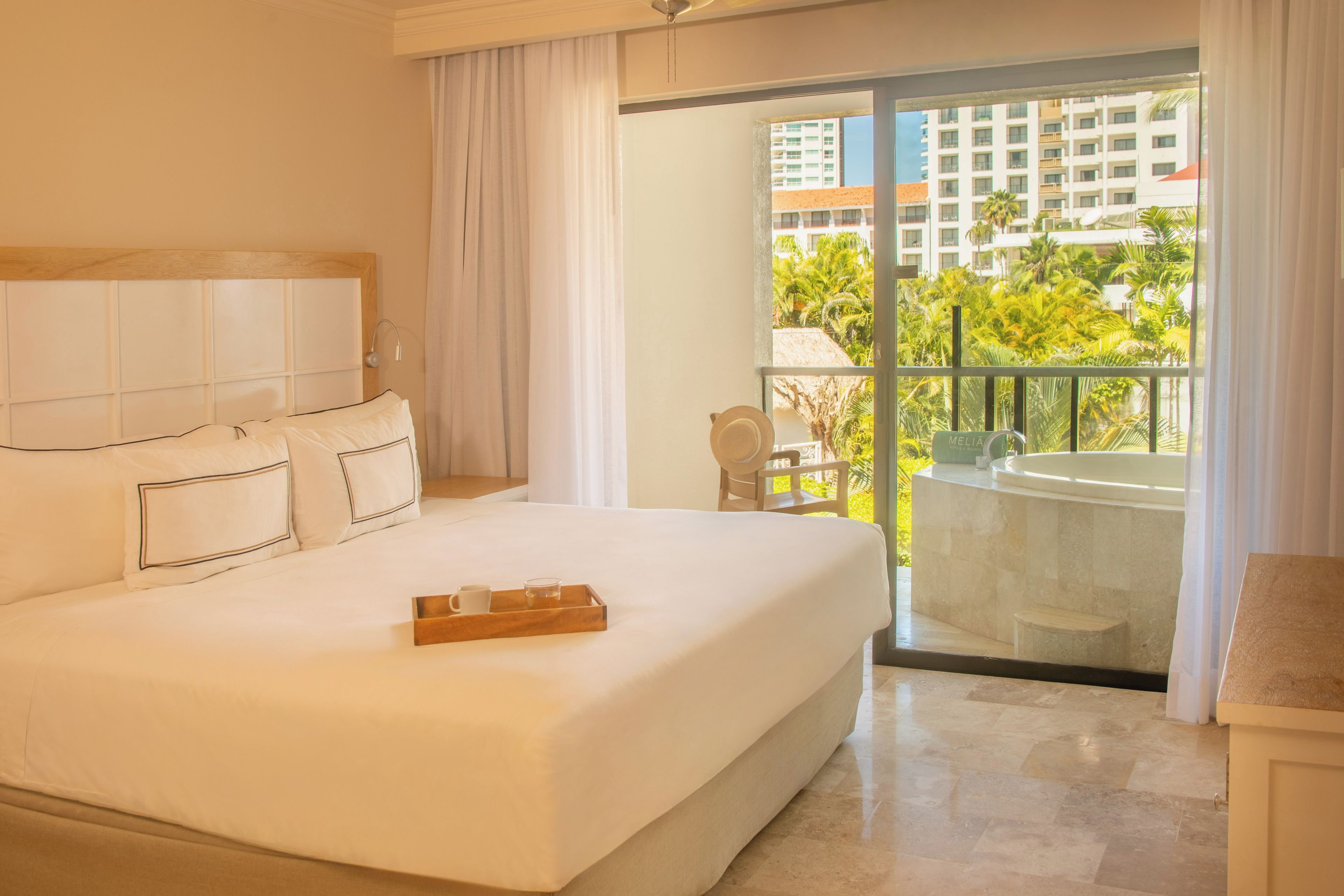 A hotel room with a large bed, a balcony with a view of palm trees and buildings, and a small table with a chair.