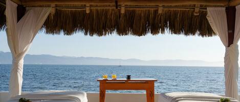 A beachside room with two beds, a small table with drinks, and a view of the sea and distant mountains.