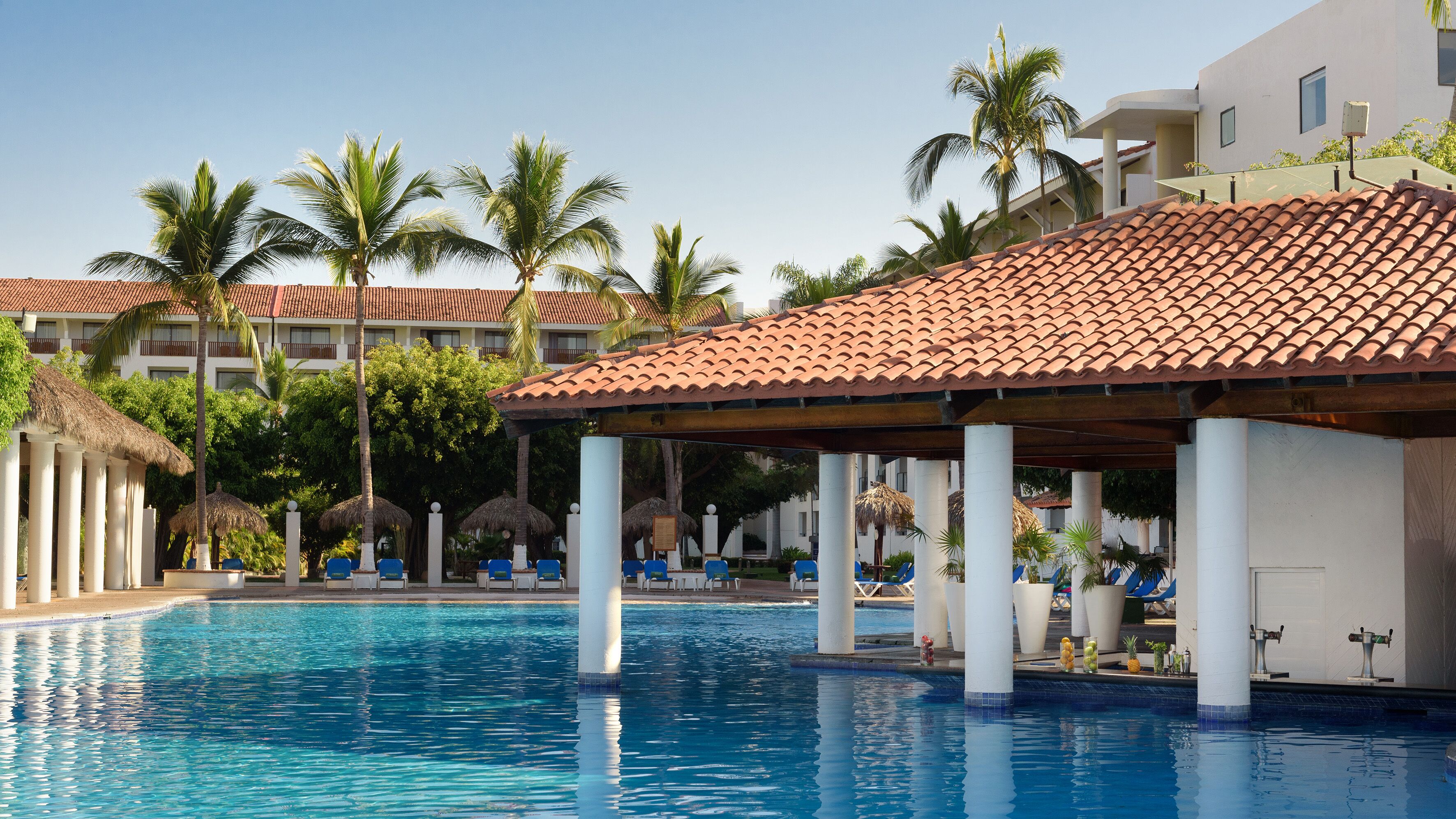Una piscina con sillas de descanso y sombrillas, rodeada de palmeras y un edificio con tejado de azulejos.