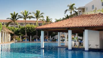 Una piscina con sillas de descanso y sombrillas, rodeada de palmeras y un edificio con tejado de azulejos.
