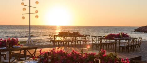 A beachside dining setup with tables and chairs arranged for an outdoor meal, overlooking the sea.