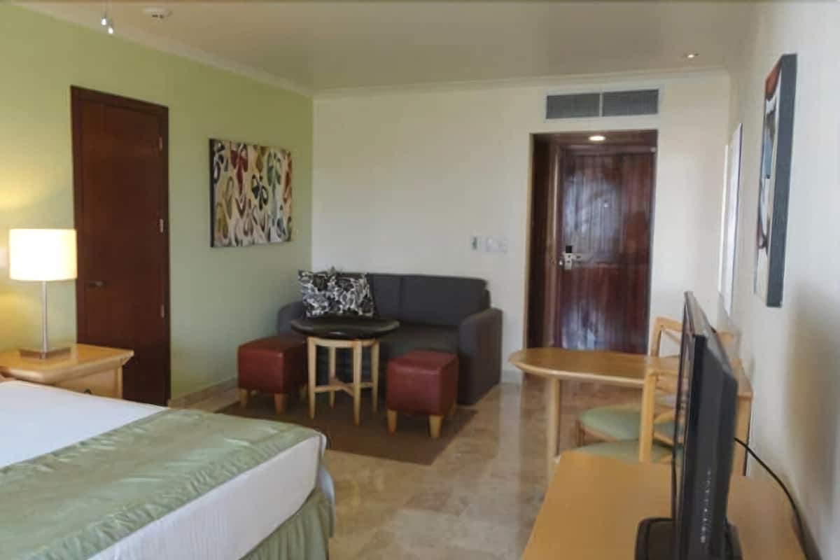A hotel room with a bed, a desk with a lamp, a sofa, a small table, and a TV.