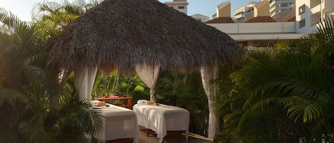 A thatched-roof outdoor seating area with two white-covered beds, surrounded by lush greenery and palm trees.