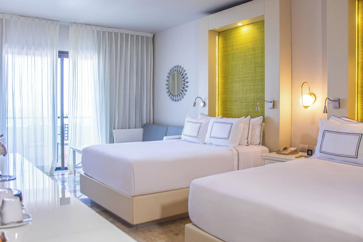 A hotel room with two beds, a large window with curtains, a mirror, and a wall-mounted light fixture.