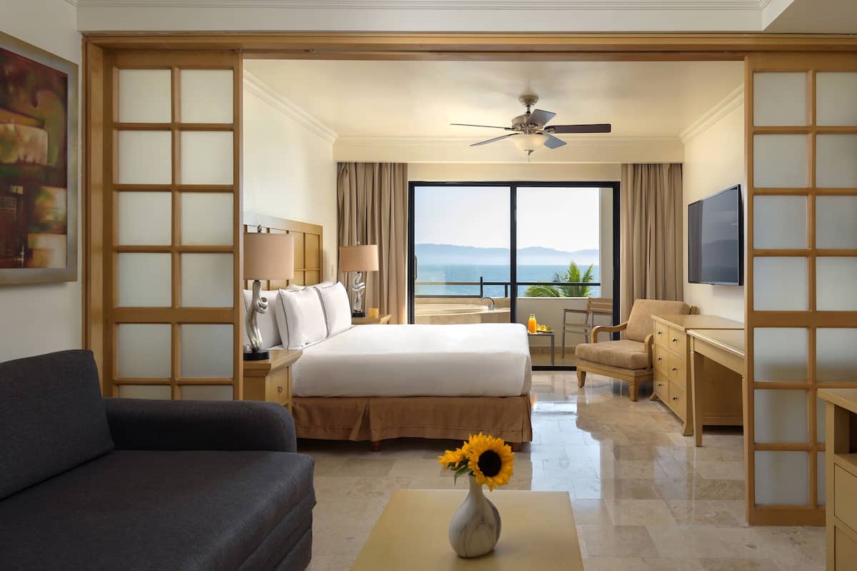 A hotel room with a bed, a desk, a chair, a sofa, a TV, and a view of the sea.