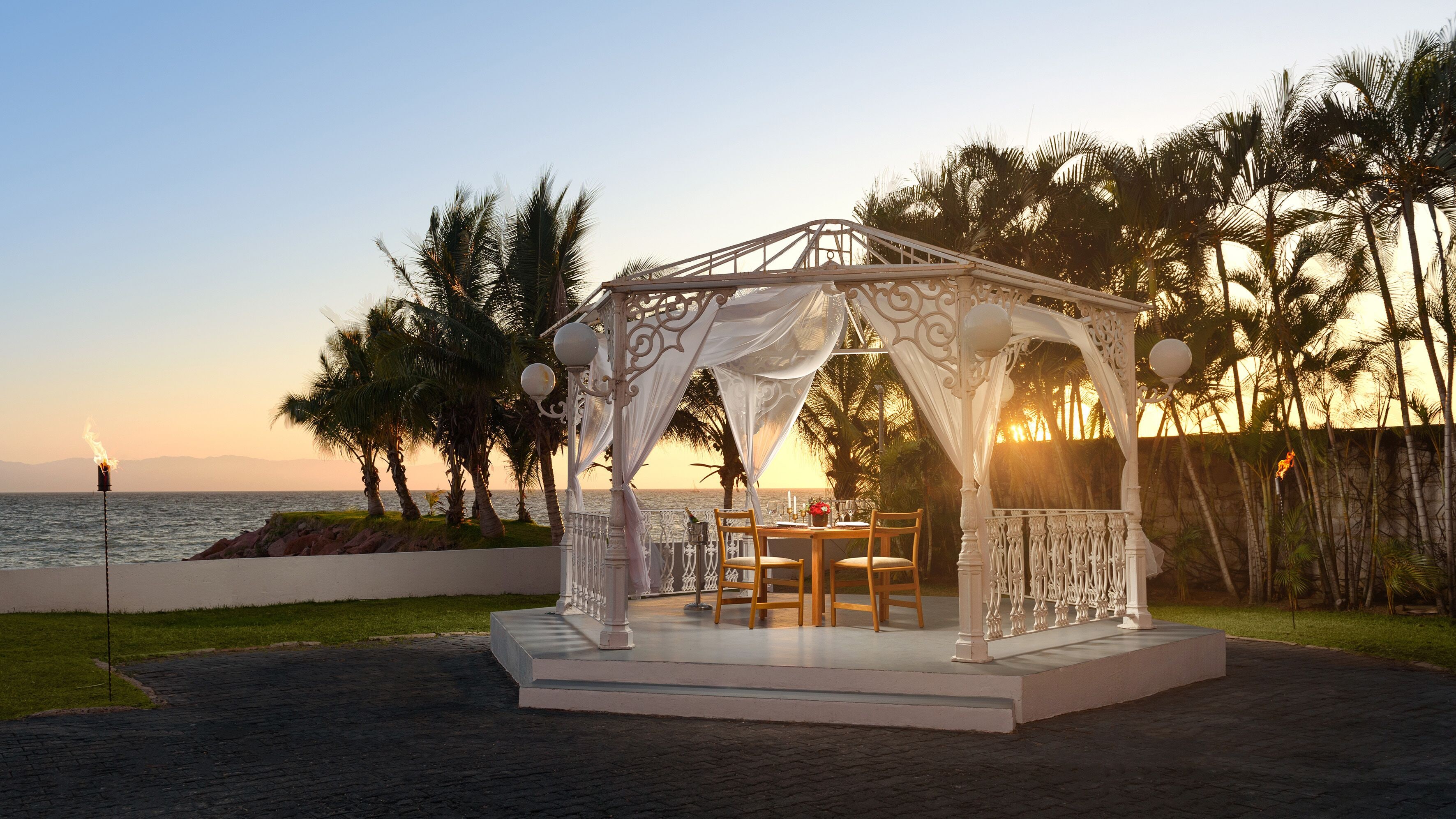 A gazebo with a table and chairs, overlooking a beach with palm trees and the ocean.