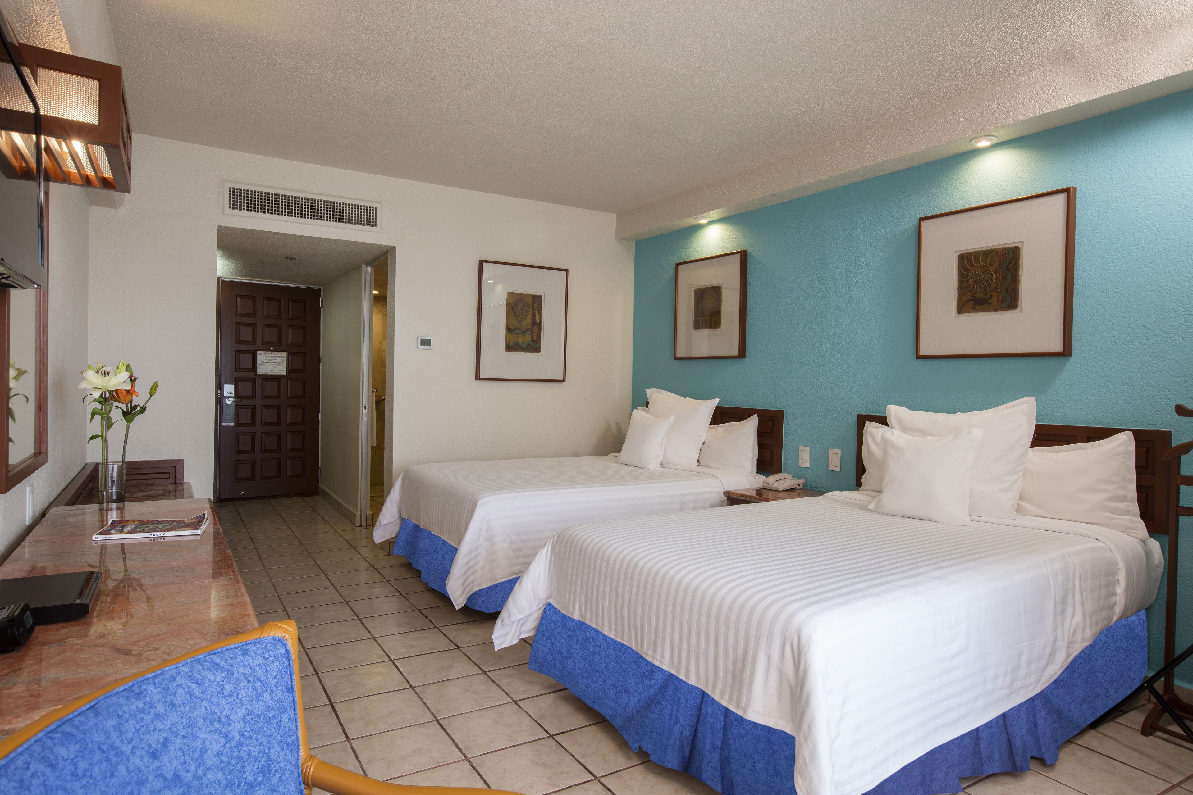 Una habitación de hotel con dos camas, un escritorio con silla, un florero con flores y cuadros en la pared.