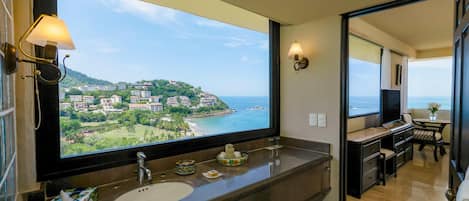 Un baño con un ventanal grande, un lavamanos, un espejo y un tocador con televisor.