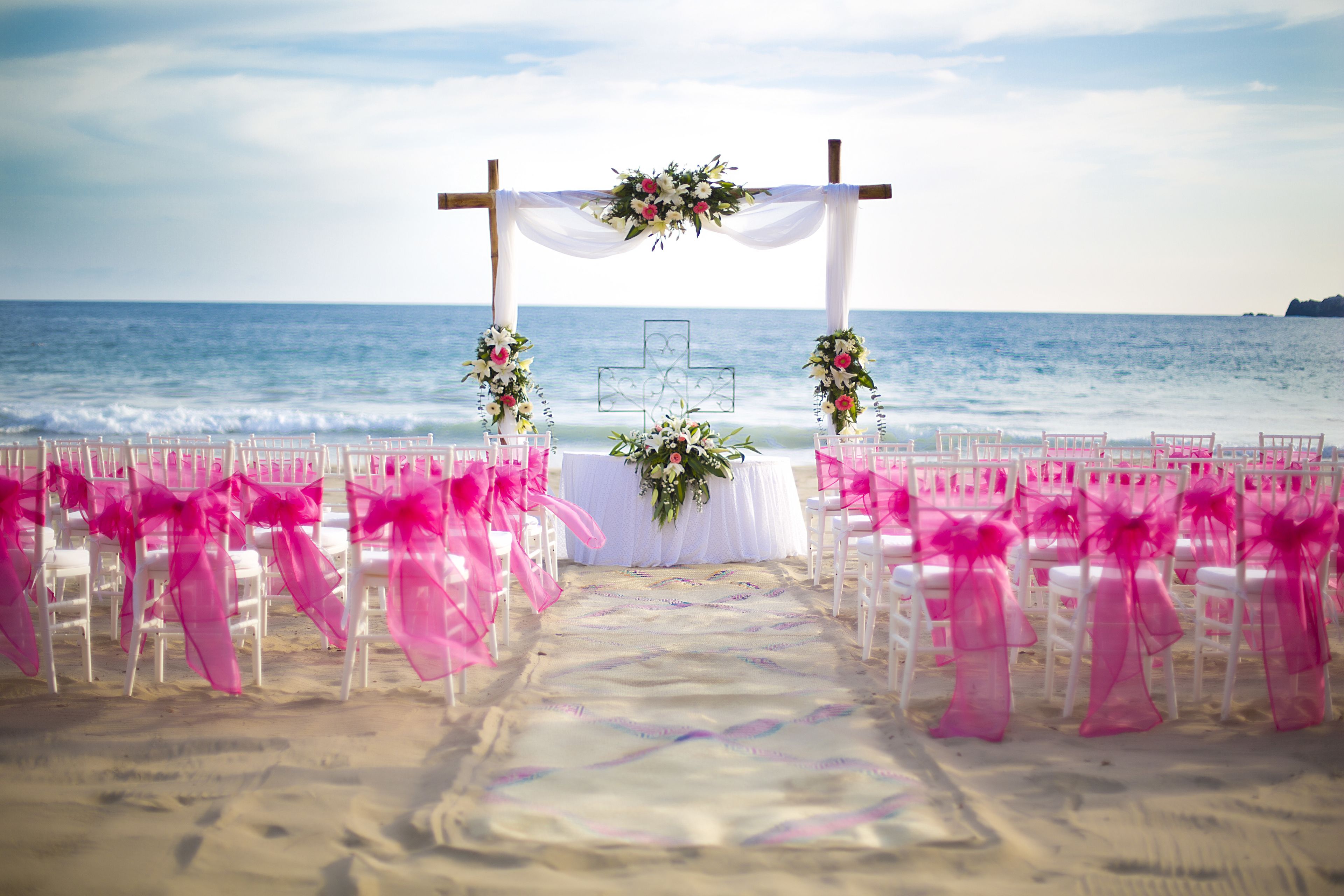 A beach wedding setup with white chairs and pink decorations.
