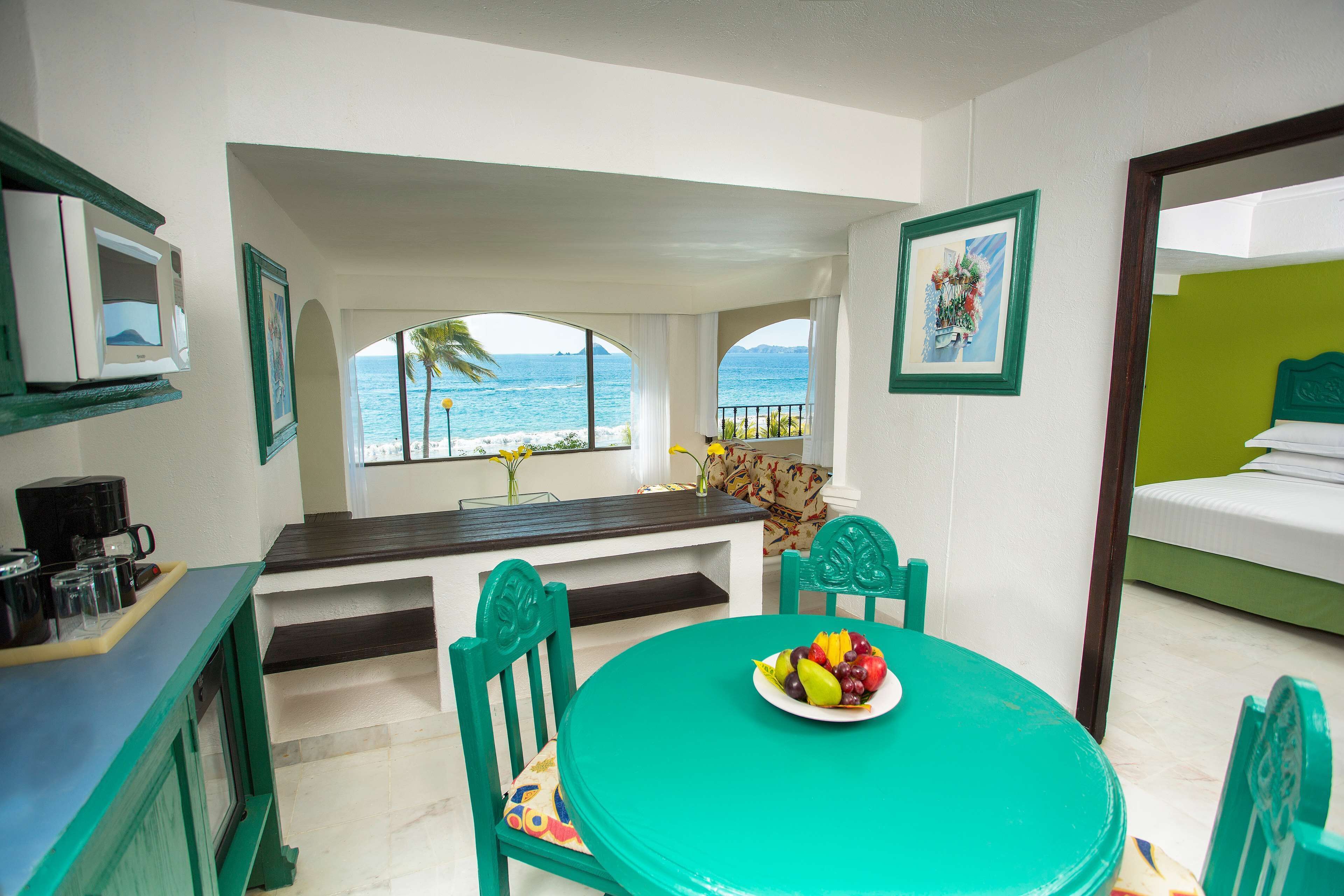 A room with a view of the ocean, a dining table with fruit, a bed, and a TV.