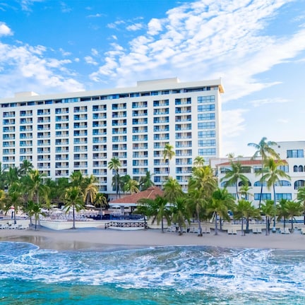 A large hotel with multiple floors and balconies, a beach with palm trees, and a view of the ocean.