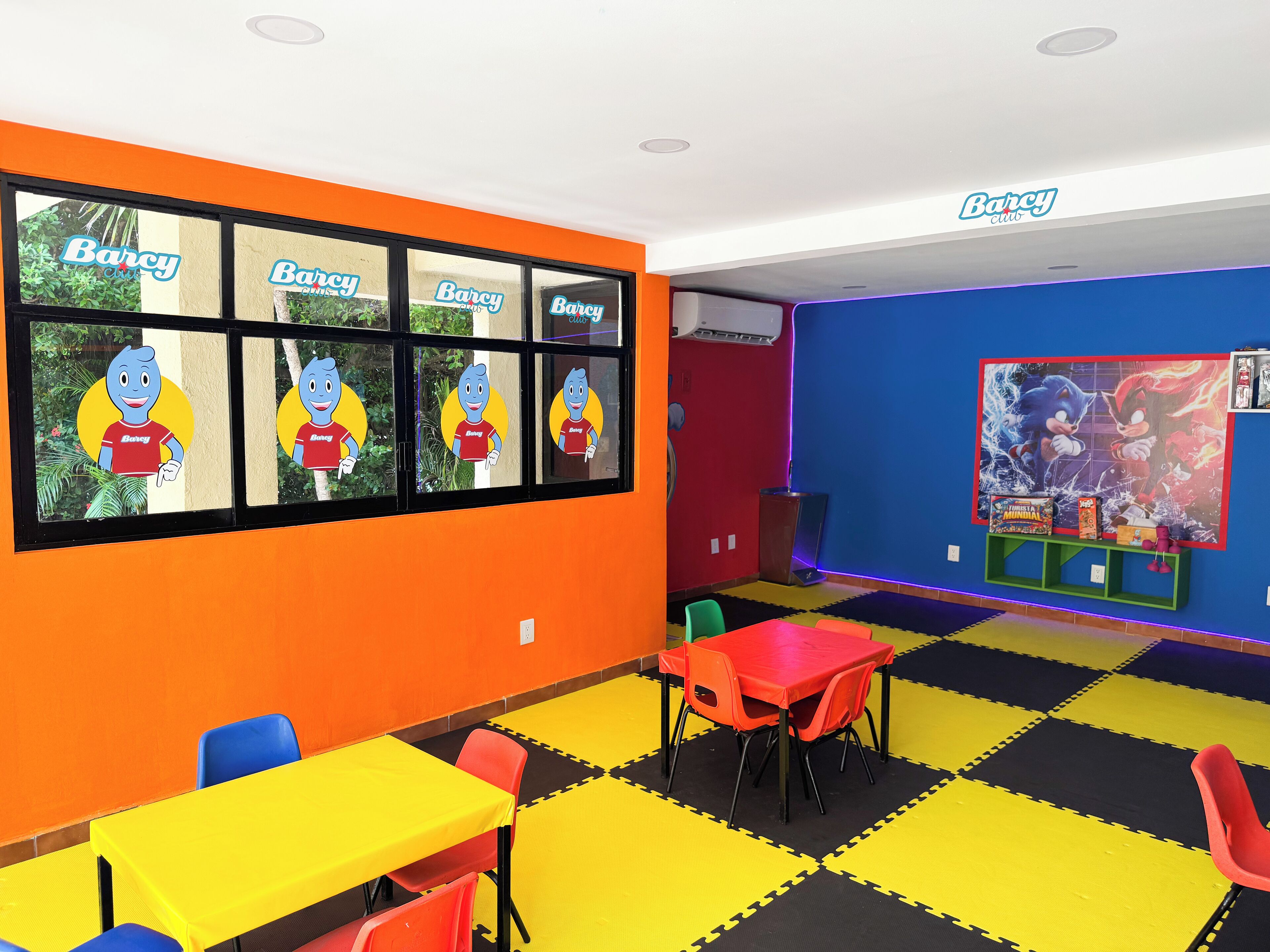 Una habitación de colores vivos con un ventanal que muestra personajes de dibujos animados, una pared azul con un póster de Sonic el Erizo y un suelo entrelazado amarillo y negro.