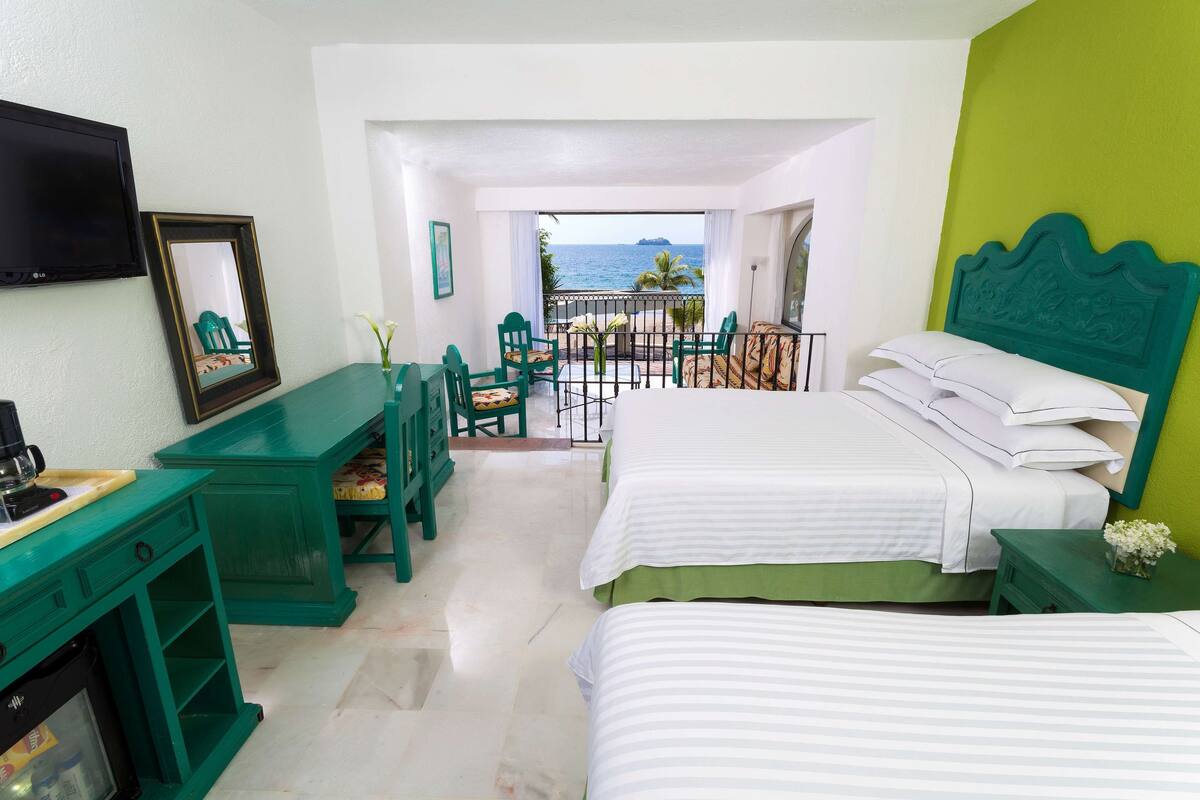 A hotel room with two beds, a TV, a desk, and a balcony with a view of the sea.