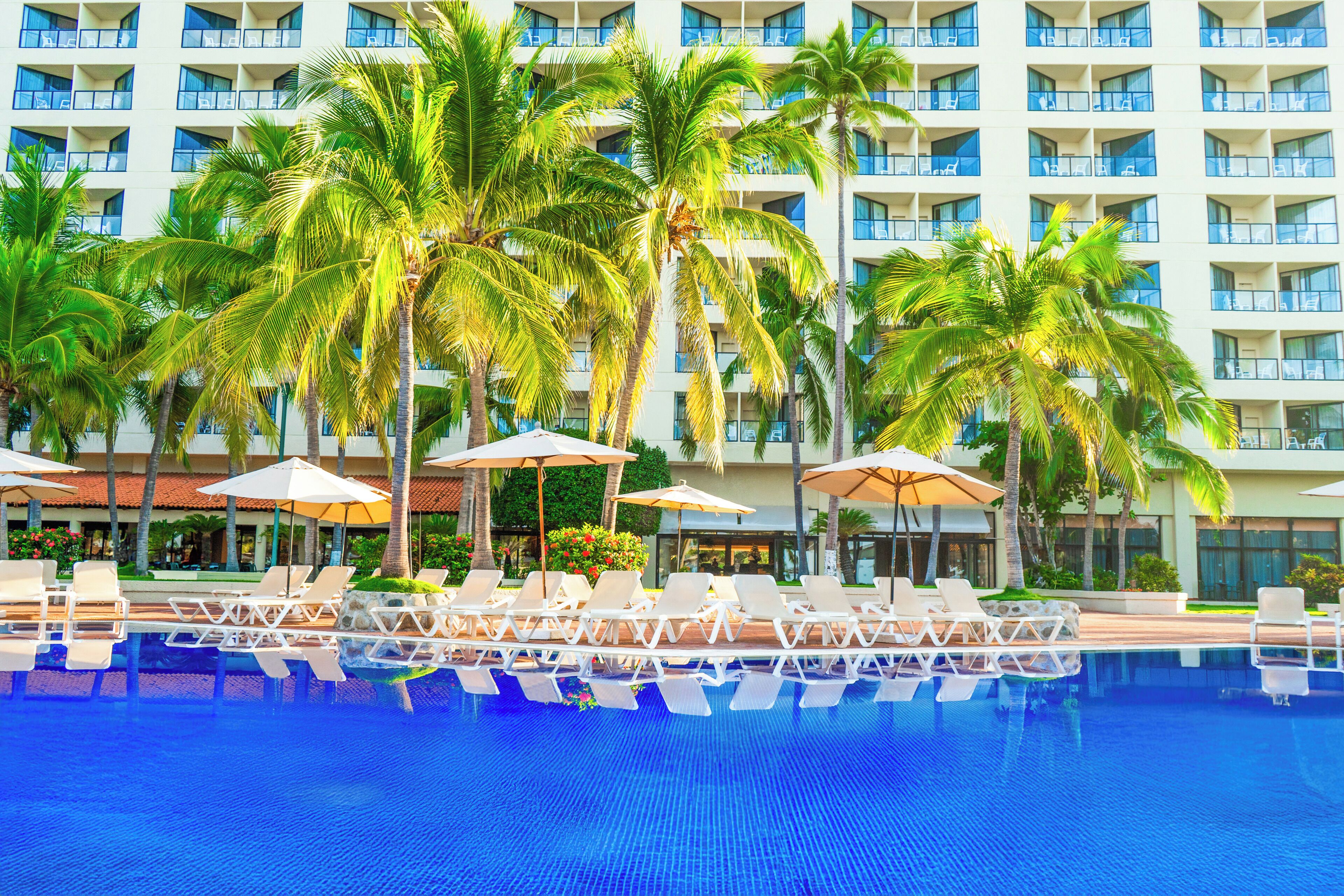 A hotel with a swimming pool, palm trees, and lounge chairs.